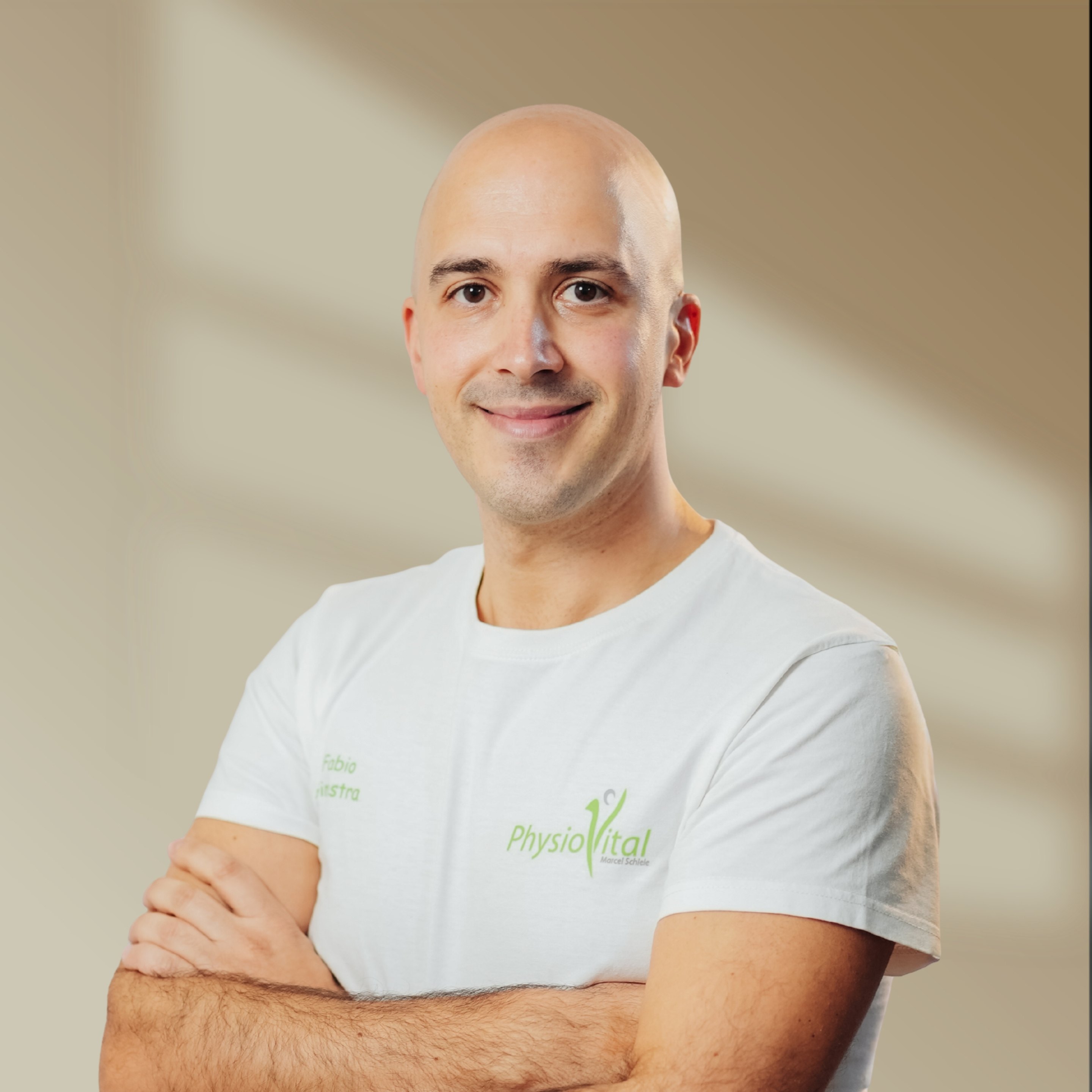 Kahlköpfiger Physiotherapeut in weißem T-Shirt mit "PhysioVital"-Logo, verschränkte Arme, lächelt.