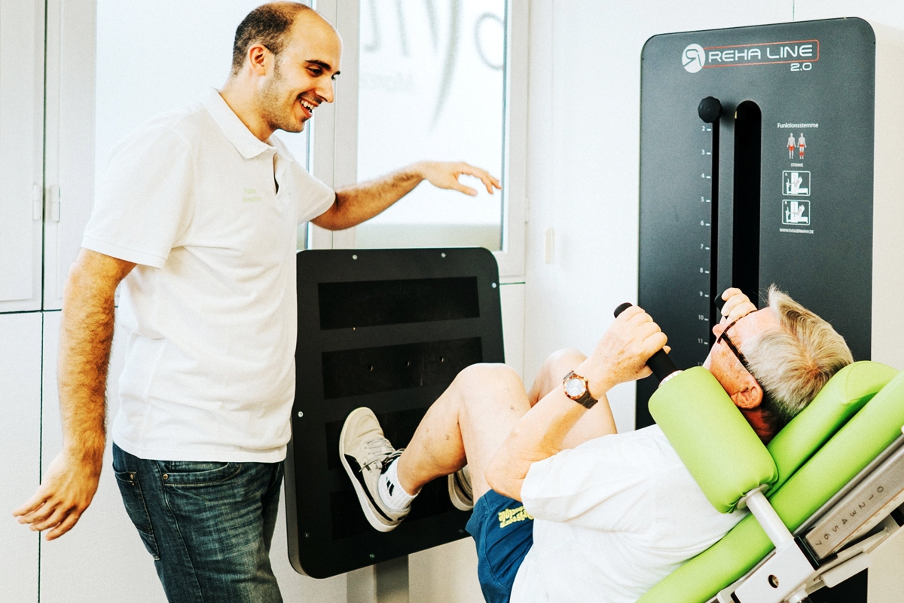 Physiotherapeut leitet älteren Patienten an einer Beinpresse mit "REHA LINE 2.0" auf dem Gerät an.