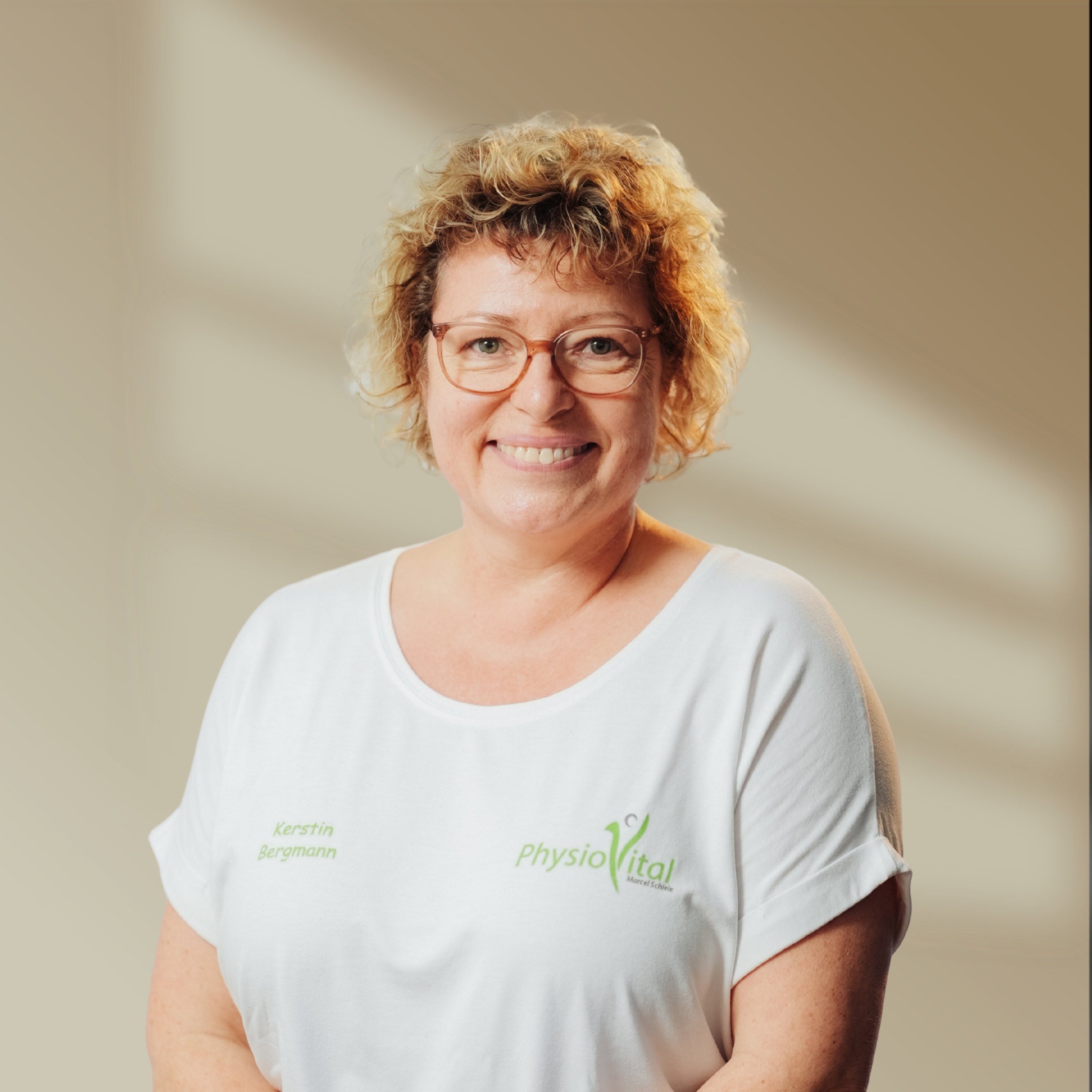 Lächelnde Frau mit Brille und blonden Locken in weißem T-Shirt mit grünem "PhysioVital"-Logo.