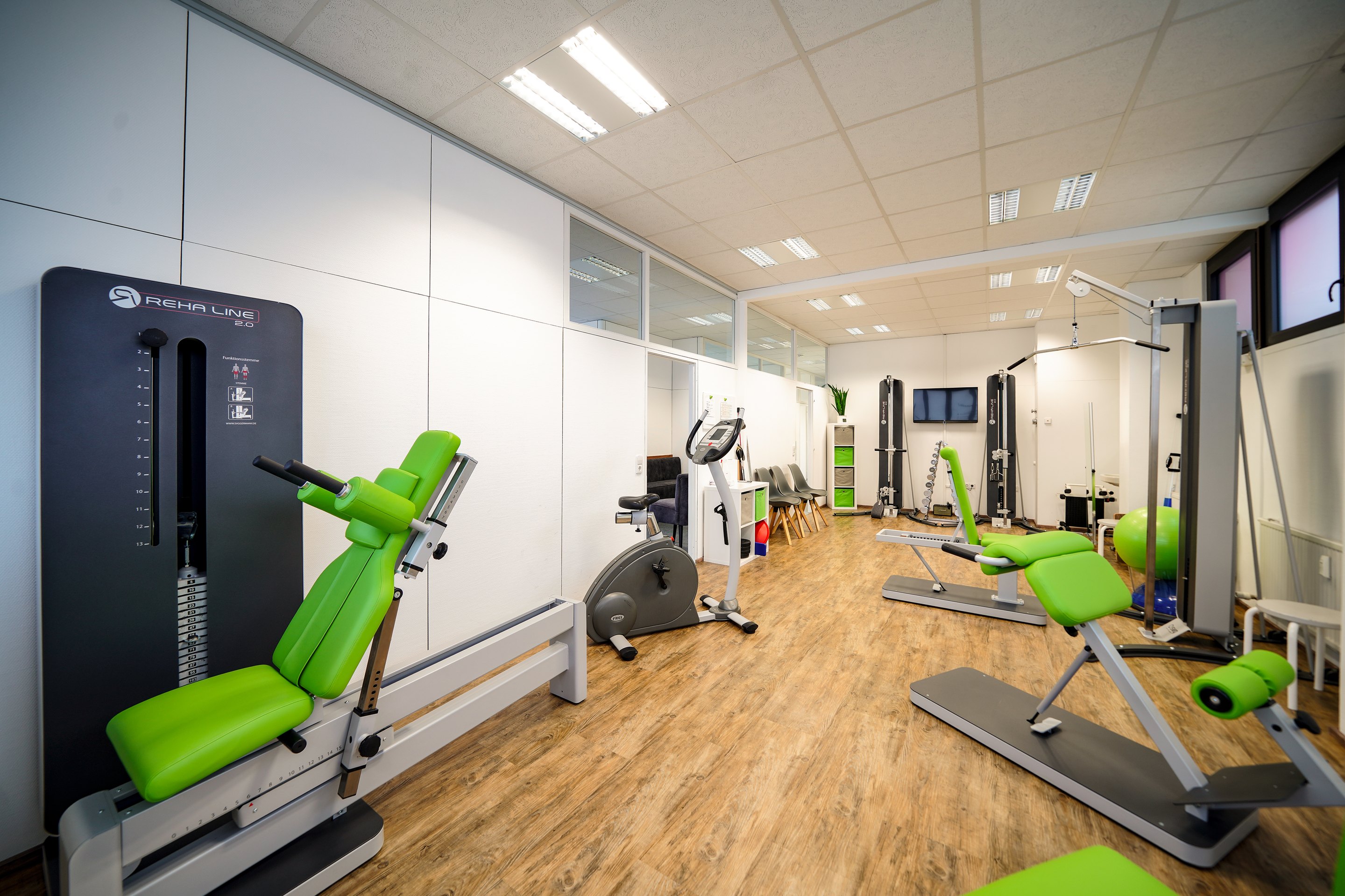 Fitnessstudio mit mehreren grünen Kraft- und Kardiogeräten auf Holzboden unter weißen Deckenleuchten.