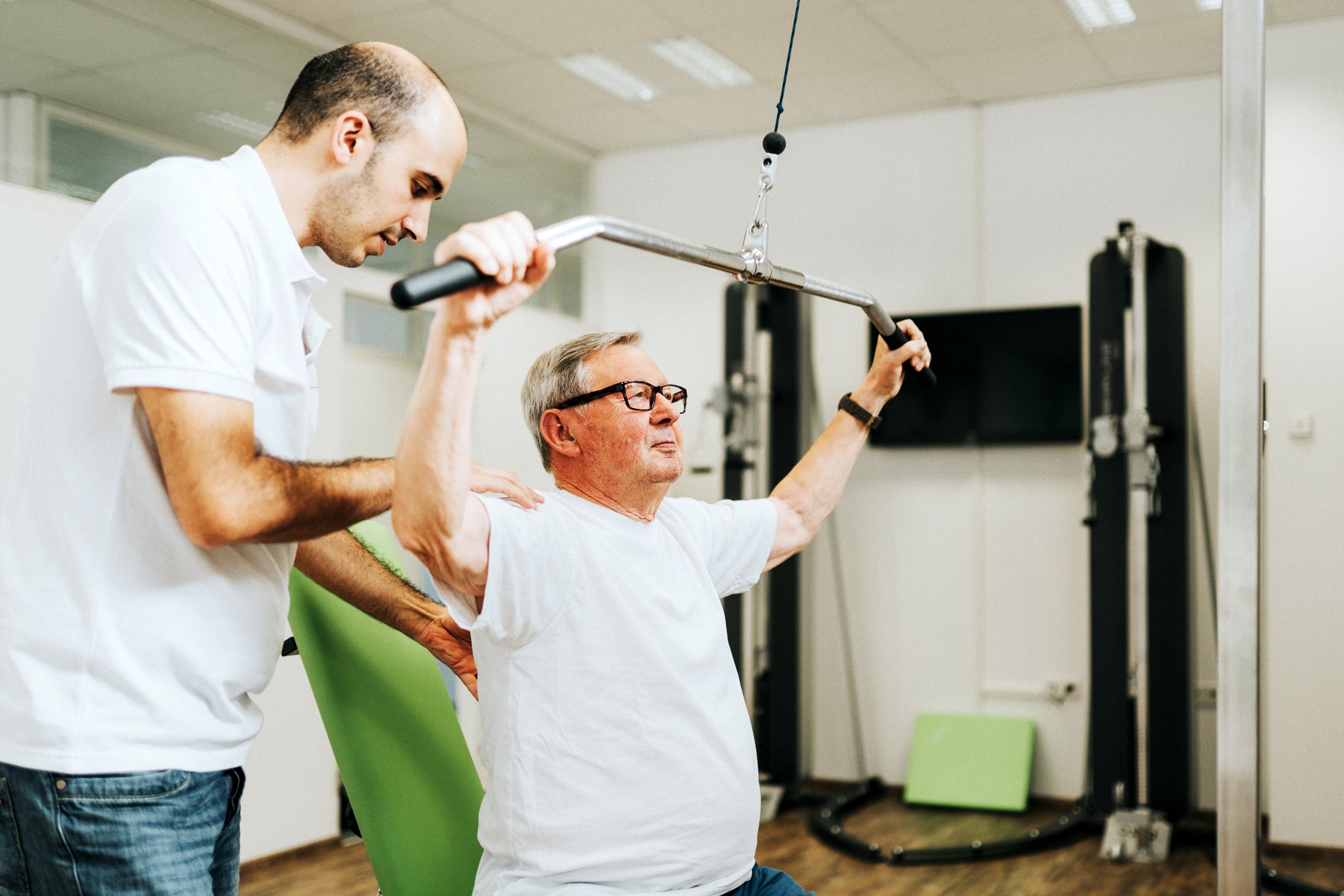 Physiotherapeut hilft einem älteren Mann beim Training an einer Latzugmaschine in einem Fitnessstudio.