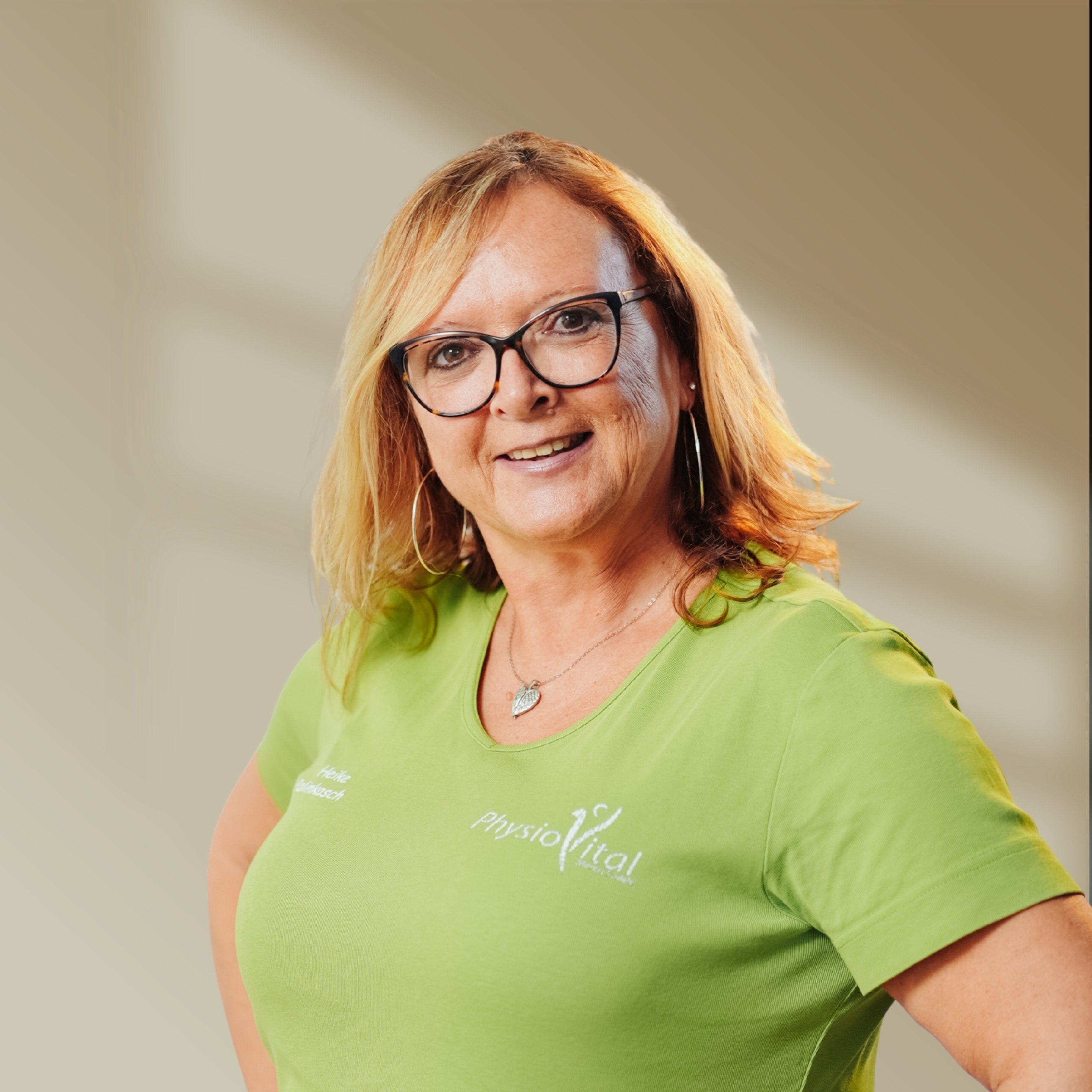 Blonde Frau mit Brille und grünem T-Shirt mit "physioVital"-Logo, lächelt zur Kamera.