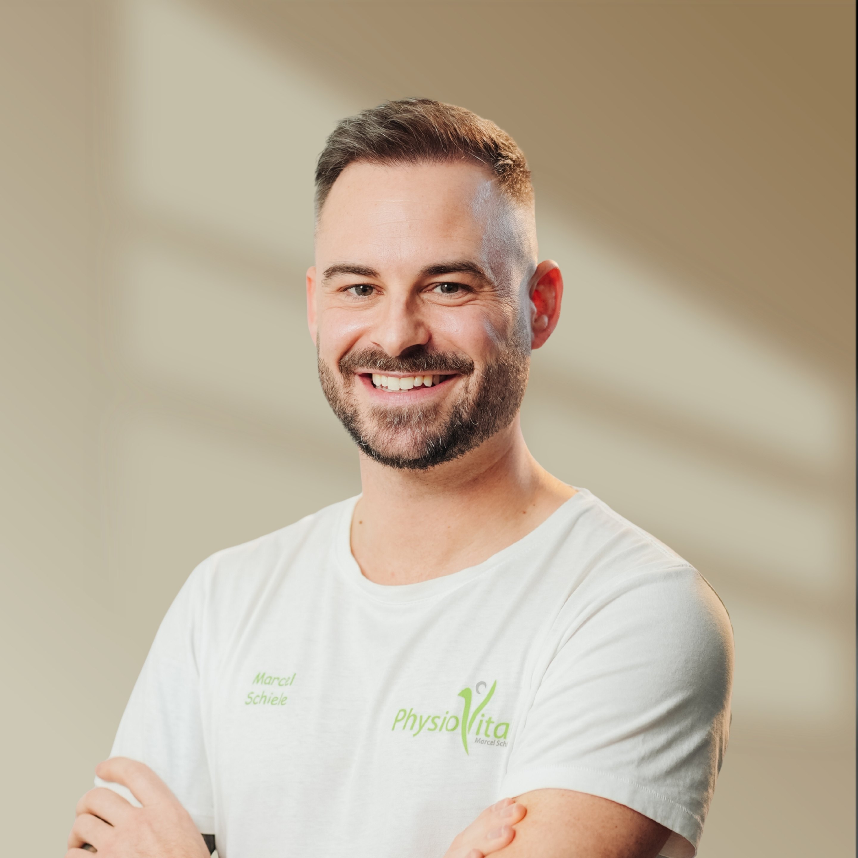 Mann mit kurzem Bart und weißem T-Shirt mit "PhysioVital"-Logo lächelt in die Kamera.