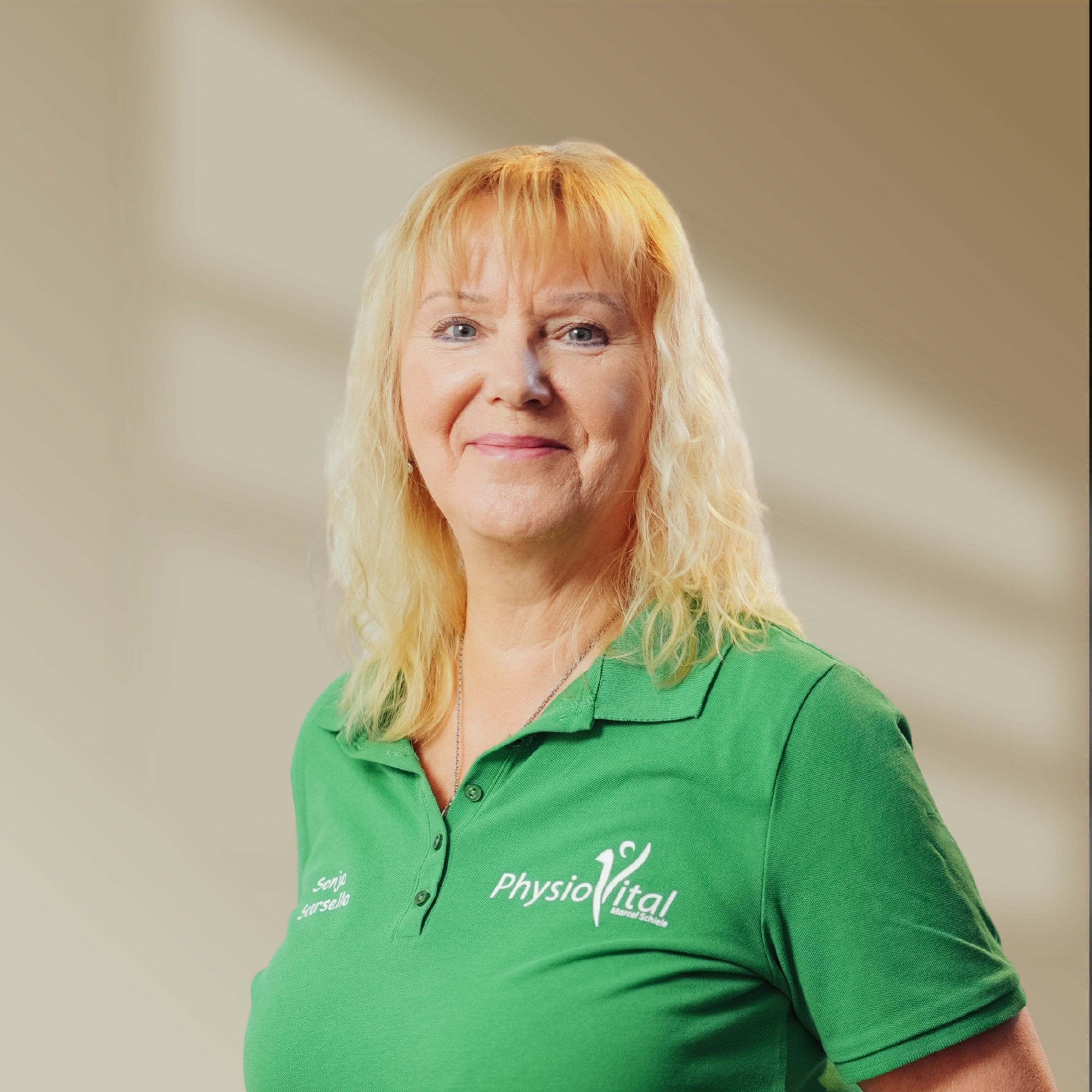 Blonde Frau in grünem Poloshirt mit "PhysioVital" Logo lächelt in die Kamera.