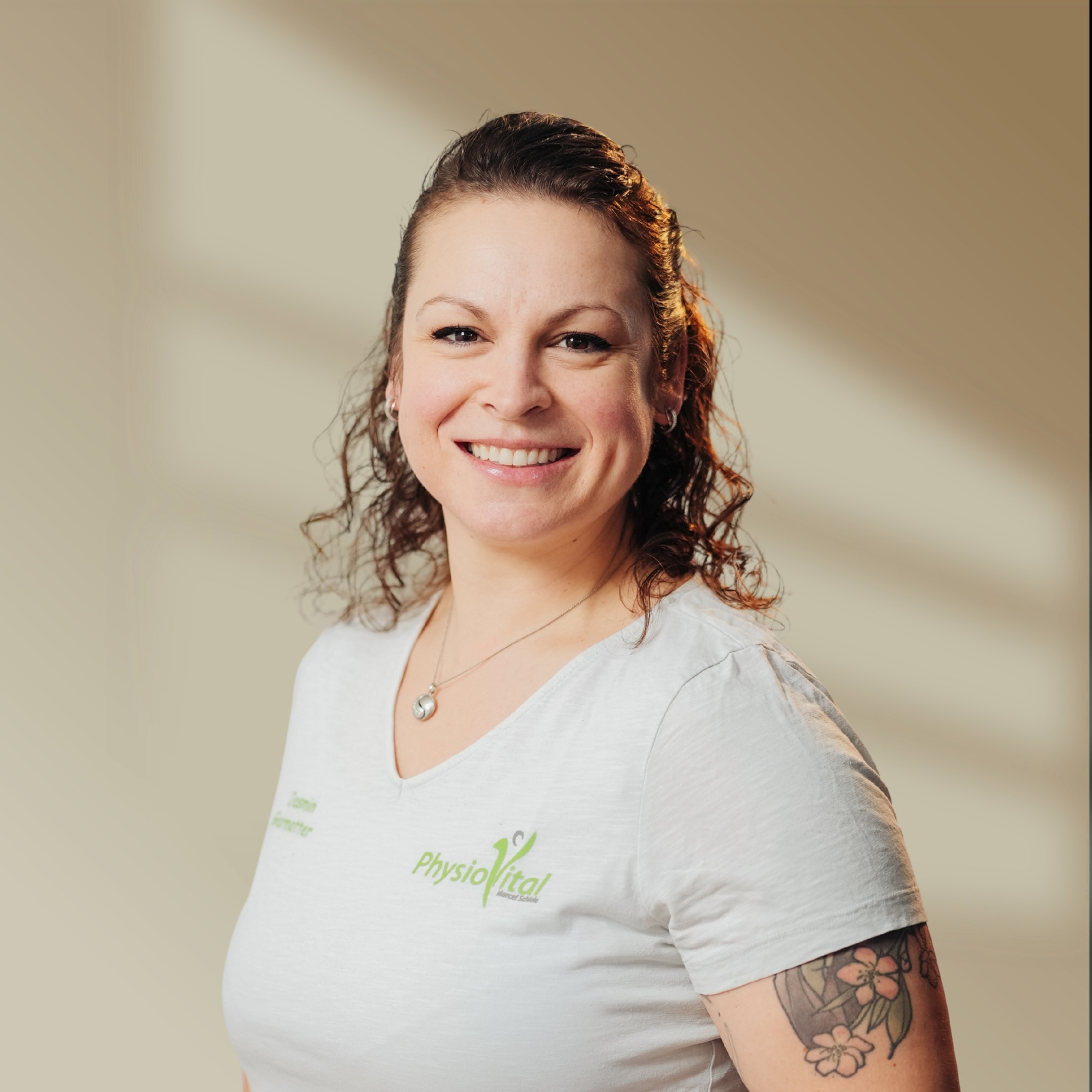 Lachende Frau mit PhysioVital T-Shirt und Blumen-Tattoo am linken Arm.
