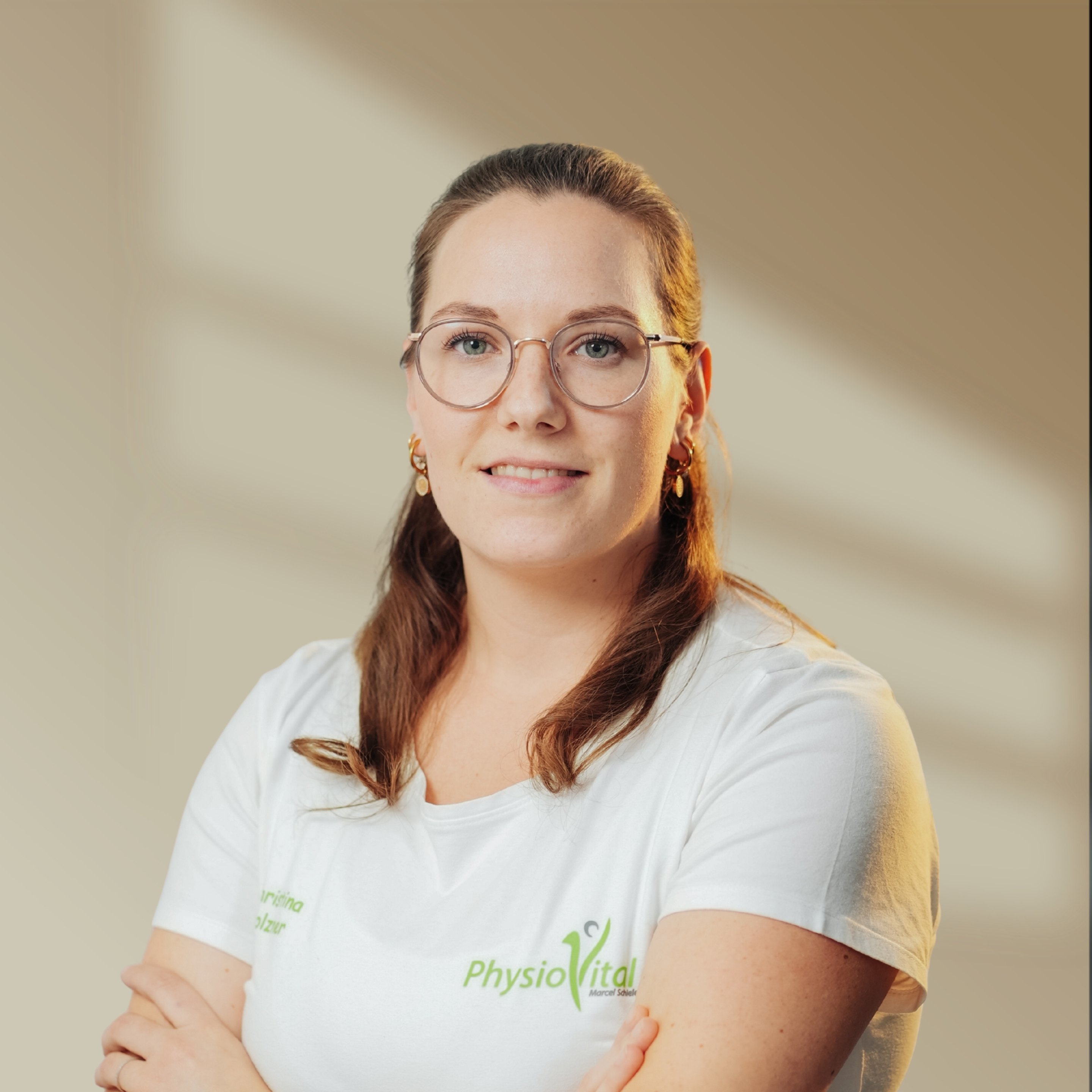 Junge Frau mit Brille und T-Shirt mit "PhysioVital"-Logo lächelt, Arme verschränkt.